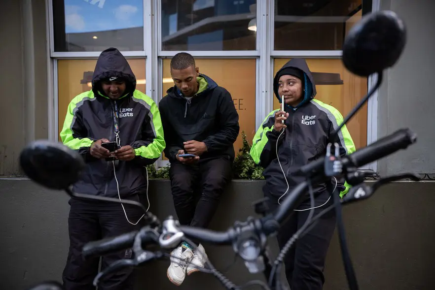 Green Riders’ (from left) Tashriq Booi, Kyle Abels and Taylor Pitcher stand outside Cavendish as they wait for order notifications to come through on their phones. Picture: David Harrison/@GroundUp News Green Riders’ (from left) Tashriq Booi, Kyle Abels and Taylor Pitcher stand outside Cavendish as they wait for order notifications to come through on their phones. Picture: David Harrison/@GroundUp News