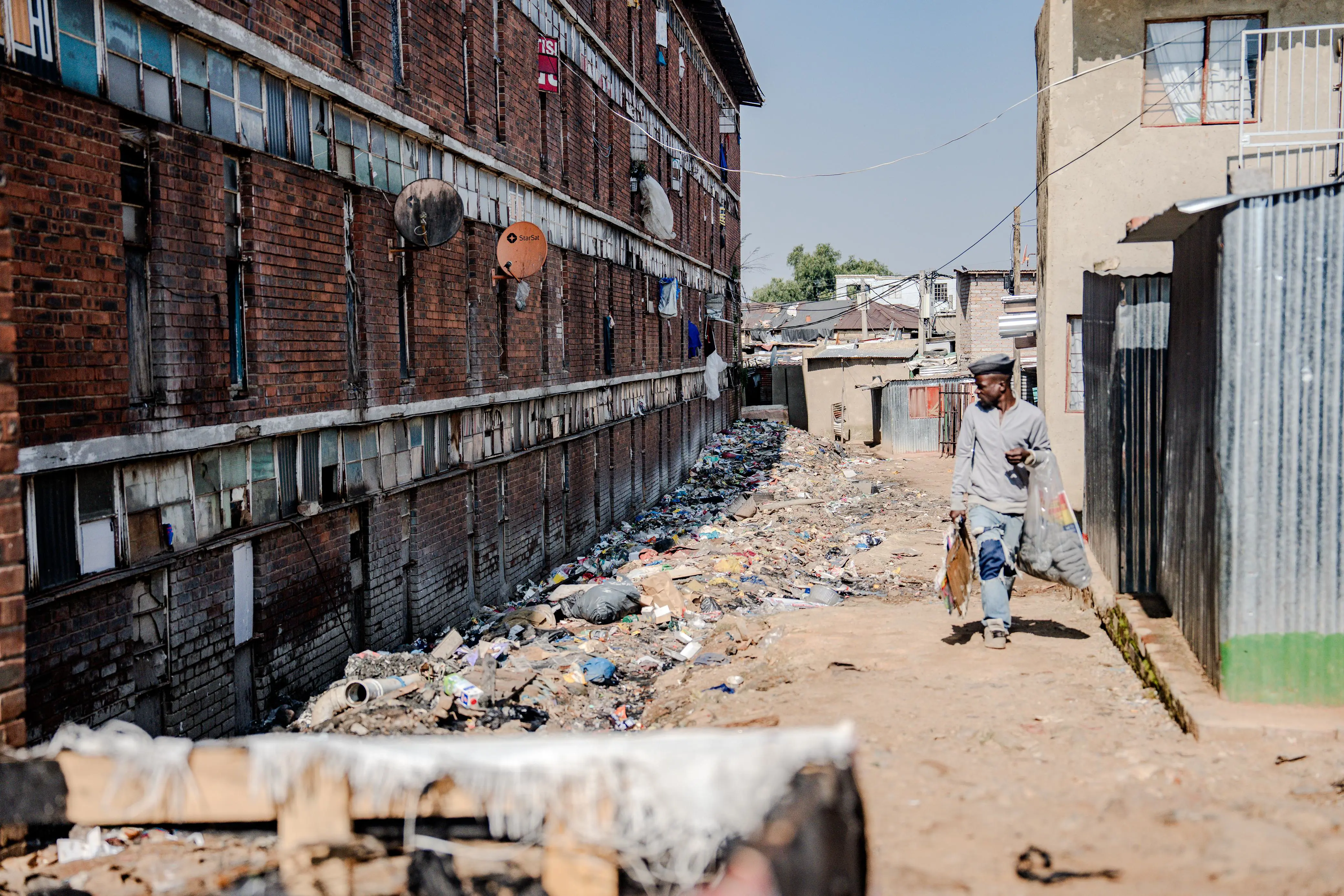 A street seller walks past the other side of the hostel, now turned into a dumping ground by residents — an everyday route shadowed by waste and survival. Photo: Sphamandla Dlamini/EWN A street seller walks past the other side of the hostel, now turned into a dumping ground by residents — an everyday route shadowed by waste and survival. Photo: Sphamandla Dlamini/EWN
